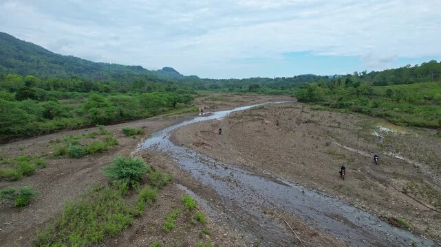Aerial drone footage of trail motorbike riders going thru a dry rocky large river in Kupang regency, Timor island, Indonesia, with forests and hills around