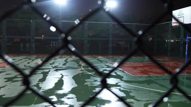 Chain-link fence close-up against a wet, blurry night basketball court with reflections.