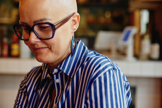 Portrait of a bold bald woman in a blue striped shirt with oversized glasses in a casual cafe setting