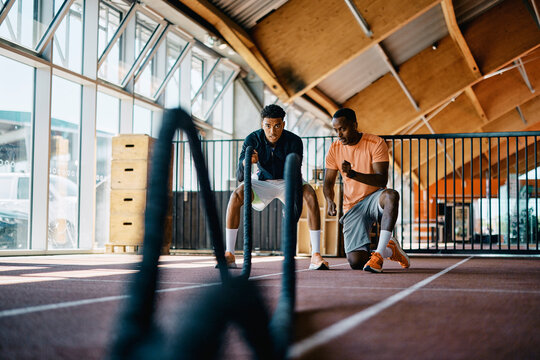 African American instructor offering encouragement to a fit young black man working out with battle ropes during an exercise session at the gym