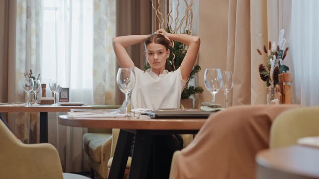 Young white woman seated at table, waiting in elegant restaurant, adjusting hair, soft daylight through curtains, empty wine glasses, warm beige tones, introspective expression, modern decor,
