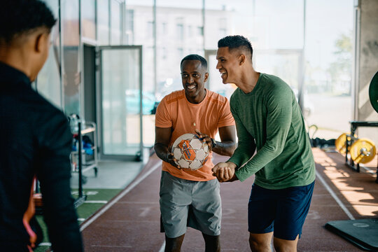 Diverse group of fit young men in sportswear laughing and tossing a ball around while warming up for a workout session together in a gym