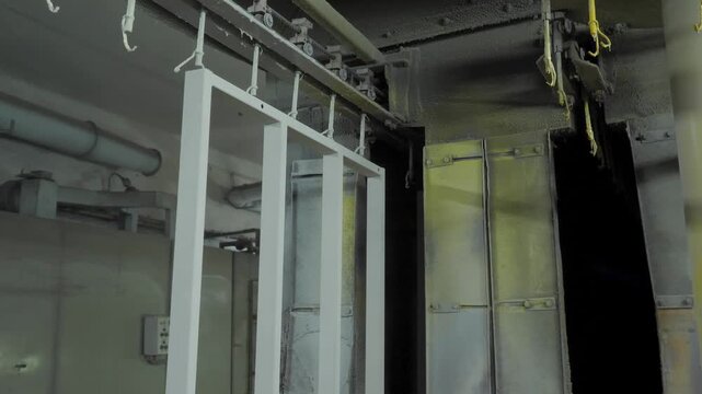 Industrial conveyor system transports white metal rectangular structures through specialized electrostatic spray booth for automated painting and surface finishing within manufacturing facility.