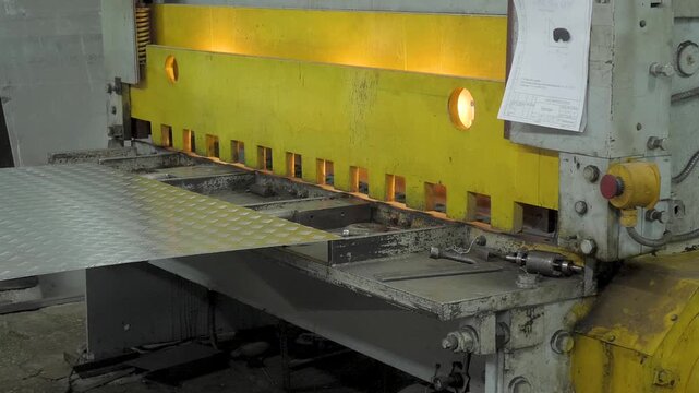 Large yellow industrial shear trims textured aluminum tread sheet into specific dimensions as part of automated metal fabrication process within manufacturing plant workshop