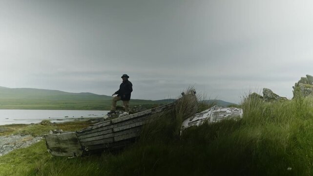 Solo male traveler enjoying a peaceful moment beside an old wooden boat on the rugged coastline of Isle of Jura near Craighouse. Cinematic Scottish island footage symbolizing adventure, reflection, fr