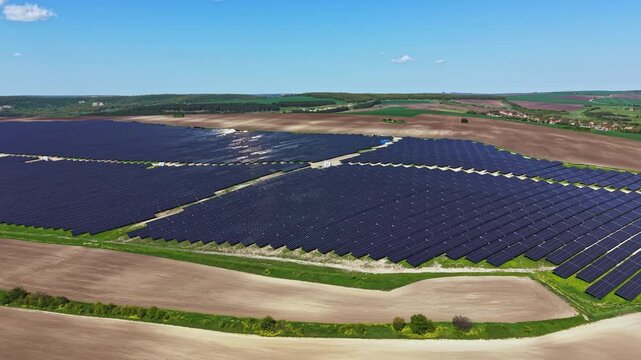 A large solar farm with many solar panels arranged in rows under a clear blue sky. The area around the farm features open fields and patches of green.