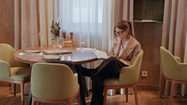 Woman jotting ideas at table. Woman seated composing notes with decorative objects nearby. Female individual dedicates time to notetaking amidst cozy cafe setting with decorative accents
