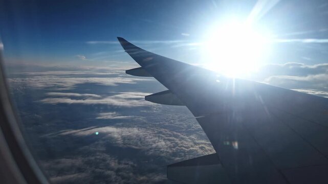 Shot taken from a passenger window seat, the aircraft wing is seen banking over a dense, textured cloud deck that extends to the horizon, illuminated by a bright, glaring sun in a deep blue sky.