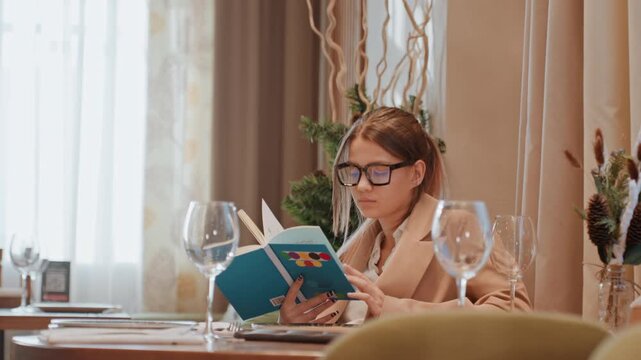 Caucasian woman reading in cozy restaurant, studying journal and notes, glasses reflecting warm light, botanical decor, peaceful posture, mindful slowliving atmosphere with elegant table setting