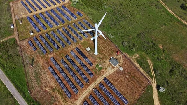 A field filled with solar panels is next to a wind turbine. The scene shows equipment for renewable energy production in a rural area. Trees dot the landscape nearby.