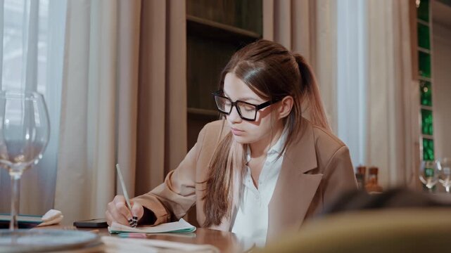 Lady notes in cozy cafe. Female professional engaged in documentation review during cafe visit. Young woman focused on annotating papers while seated in warm and inviting coffee shop