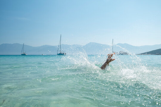 Young boy's arm splashing in clear turquoise sea of iconic Saleccia beach, with sailboats and mountains, evoking summer travel and relaxation at Saleccia and Lotu beach, Corsica.