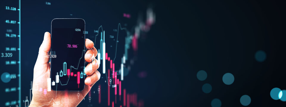 Hand holding smartphone displaying glowing financial chart against dark digital background showing market data and investment concept.