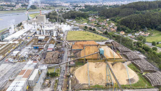 Aerial view of Biofabrica de Ence with large wood chip piles, industrial structures, and smoking chimneys near lush green hills Pontevedra, Pontevedra, Spain.