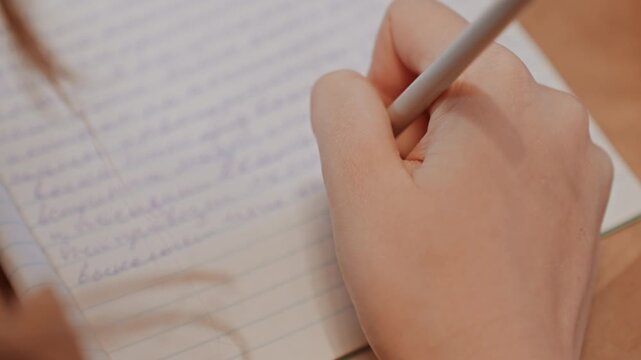 Closeup hands writing on notebook, student taking notes at desk, pencil tracing cursive on lined paper, warm classroom lighting, focused study atmosphere with visible sweater sleeve and partial hair