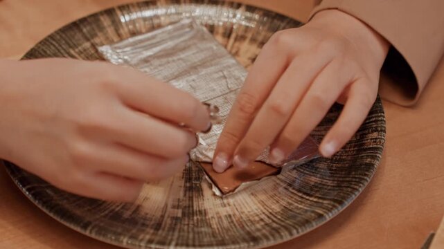 Closeup hands unwrapping chocolate bar on plate, home baker carefully peels foil to reveal glossy milk chocolate, decorative ceramic plate, warm amber lighting, cozy kitchen atmosphere, manicure