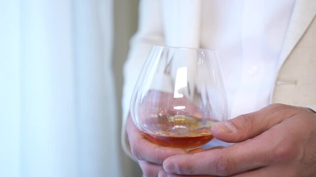 Elegant person wearing a light-colored suit and holding a glass of amber cognac. Wedding ceremony. Groom holding a glass of alcohol. Ideal for themes of business success, relaxation, fine spirits, and