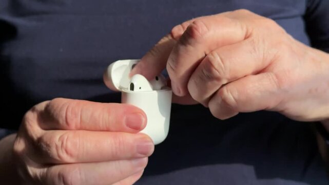 Close-up woman holding white earbud case, gently removing single earbud for listening warm sunlight highlights textured hands