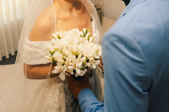 Couple stands together in formal attire with a bouquet in bright room. couple holds each other close in a well-lit room. bride wears a white dress and holds a  flowers. 