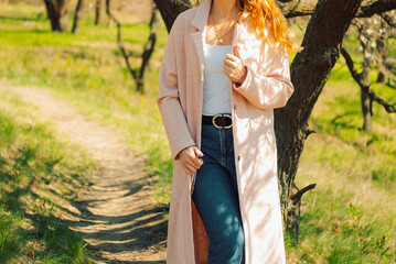 Woman stands outside wearing  light coat and jeans while holding onto her coat in a green field during daytime. outdoors in green field. She wears  light coat over  tank top and jeans.   © SValeriia
