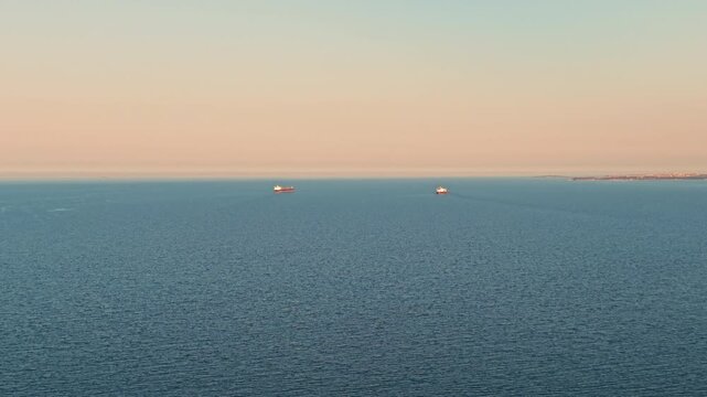 Two ships navigate across the vast sea, visible on the horizon. The water is smooth and reflects the colors of the early morning light. The sky is clear, creating a serene background.