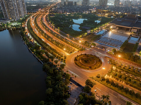 Blue hour aerial view of illuminated roundabout and boulevard beside lakeside residential district in western Hanoi.