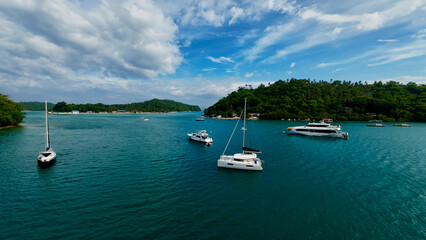 Luxury yachts and sailboats anchored in a tropical island bay. Aerial seascape with turquoise water, green hills and dramatic clouds over the ocean. © Houston