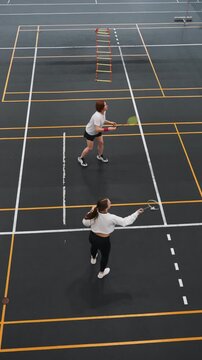 Two players indoor badminton doubles rally overhead gym court lines fast footwork and coordinated teamwork during intense backcourt exchanges shuttlecock flight visible training ladder in background