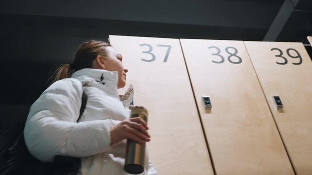 Woman entering locker room carrying bag, opening numbered locker and holding bottle, wearing white puffer jacket with long ponytail, modern wooden lockers with visible numbers, focused expression,