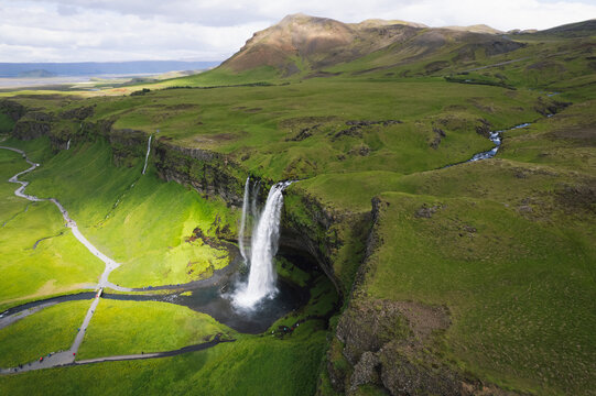 Aerial view of Seljalandsfoss waterfall cascading over a cliff into a pool surrounded by lush green hills and walking paths Rangarthing eystra, Iceland.