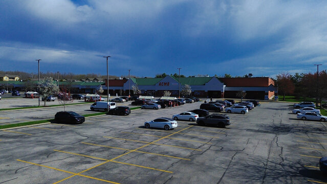 Elevated drone view reveals the ACME Markets grocery store facade bordered by blooming spring trees and parked customer vehicles across the expansive asphalt lot. Delaware