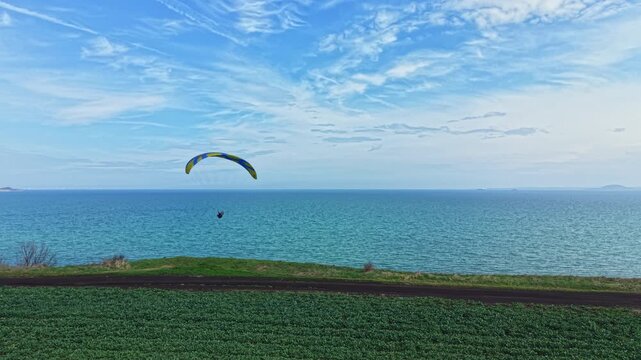 A person is paragliding over the ocean with calm waters below. Green grass lines the shore while clouds are visible in the blue sky. The scene shows the joy of flight.