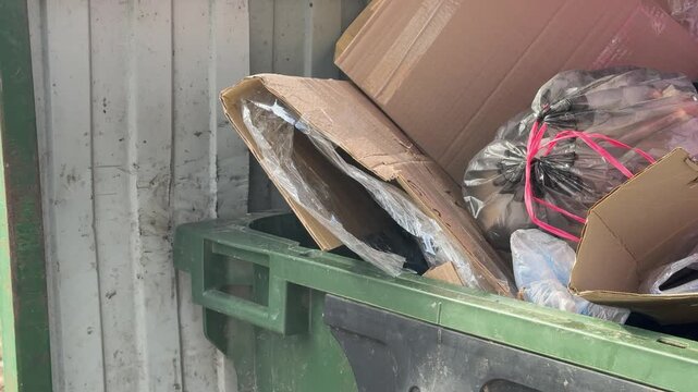 Close-up overflowing green dumpster loaded cardboard and construction debris including insulation, taped bags, crushed packaging