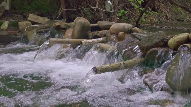 A serene close-up of a handmade stone water cascade in a lush green forest. Clear water flows through metal pipes into a bubbling stream, creating a tranquil and natural atmosphere. Ideal for wellnes