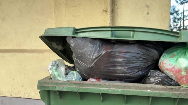 Close-up overflowing green dumpster loaded cardboard and construction debris including insulation, taped bags, crushed packaging