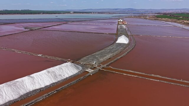 Salt flats stretch across the land with red water reflecting the sky. Salt piles sit on the sides of the paths leading through the flats. Fields grow beyond the water.