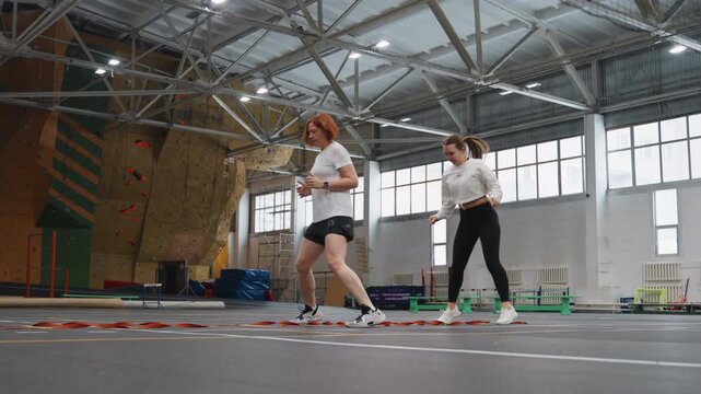 Badminton footwork drills indoor sports hall coach guides athlete through stepping patterns on marked court with high ceiling and climbing wall in background, emphasis on quick lateral steps, balance,