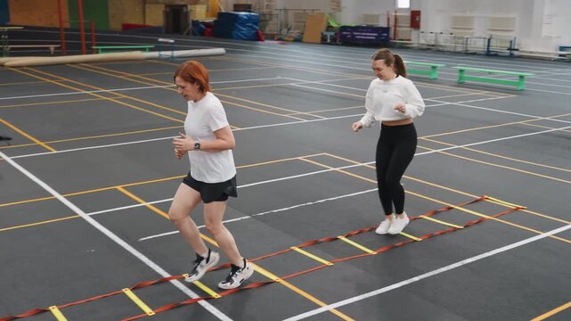 Two white women practicing badminton footwork ladder drills on indoor court, coach demonstrating quick steps while teammate follows, marked court lines and colored cones, focused expressions,