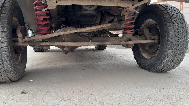 Close-up crossover suspension undercarriage view dusty asphalt gravel ground, red coil springs and muddy axle visible, worn tires
