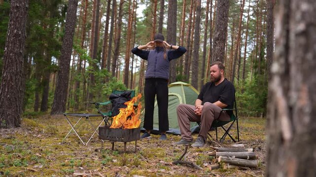 Young woman doing relaxing stretching exercises next to a campfire while a man sits nearby in a forest campsite. Active lifestyle and outdoor recreation with a tent and bonfire in the background