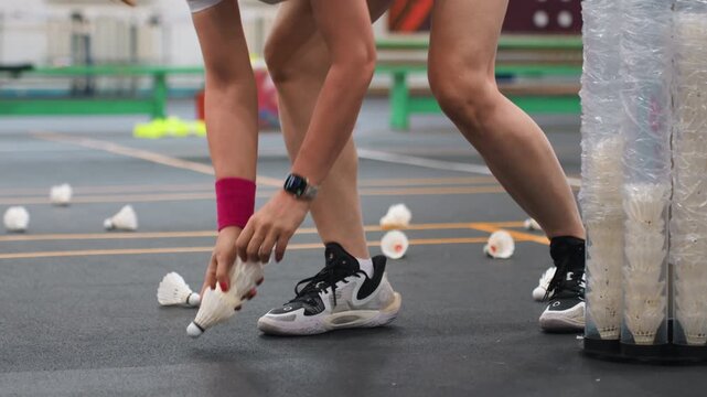 Player collecting shuttlecocks on indoor court, hands and legs visible as athlete picks scattered shuttles from floor and plastic tube, pink wristband, black shorts, white sneakers, focused training