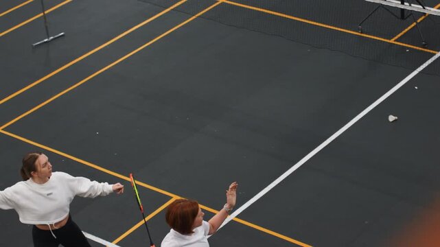 Two women contesting fast badminton rally indoor court, powerful overhead smashes and athletic jump reach, rapid footwork and quick returns, shuttlecock flight across net, focused expressions