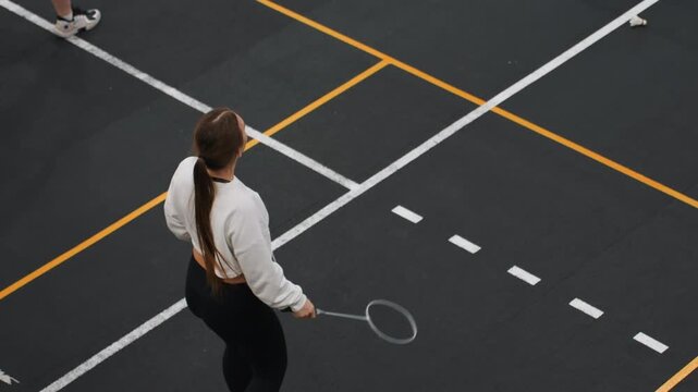 Overhead court solo woman practicing badminton serve and footwork, rhythmic repetitions across marked lanes, controlled swing and eye tracking, focused training session in indoor gym