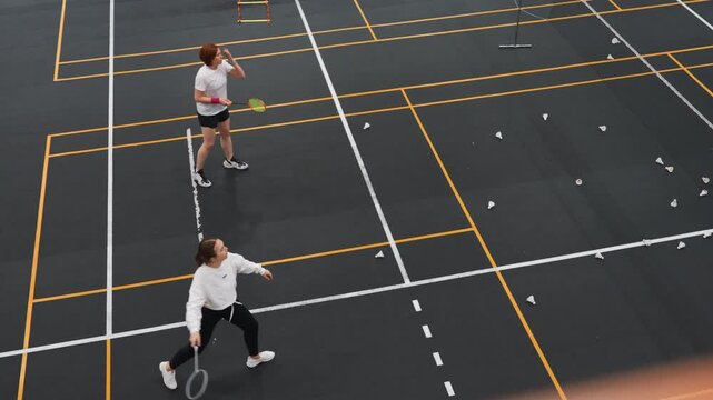 Indoor badminton rally with women players overhead court view, fast volleys and smashes, tactical footwork, coach observing, shuttlecock flight, marked court lines, gym lighting, highenergy training