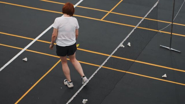White woman collecting shuttlecocks by net during repetitive feed drill on indoor court with yellow markings low reach pickups, precise placement, quick recovery steps and ladder drill in background