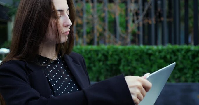 Caucasian woman outdoor caf holding tablet, polka dot blouse and blazer, confident smile as she scrolls presentation on device, urban garden backdrop, natural warm light and relaxed professional