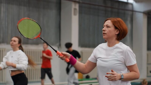 White female player practicing badminton rally, redhaired amateur concentrating on serve and dink in indoor gym, wearing white tee and pink wristband, holding bright racket, focused expression,