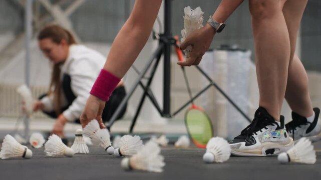 Shuttlecocks scattered on indoor court, player kneels to gather white feathered shuttlecocks near sneakers and racket assistant adjusts tripod while coach observes drill closeup on hands and feet