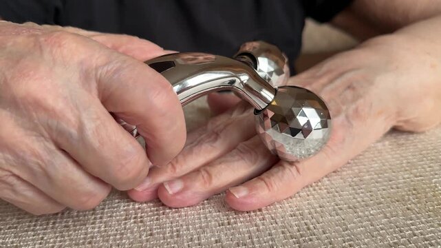 Elderly hands massaged with roller close-up silver two ball massager gliding over wrinkled forearm on textured fabric gentle