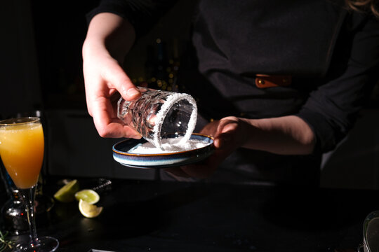 Bartender woman making salt rim on cocktail glass using small plate. Professional preparation for Margarita drink at bar counter.